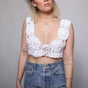 May include: White crochet crop top with a tie detail in the front. The top is made of a delicate, open-weave fabric and has a relaxed, bohemian style.