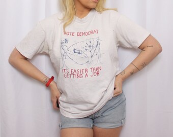 May include: White t-shirt with blue text that reads "Vote Democrat. It's easier than getting a job." The shirt features a cartoon illustration of two people in a hammock.