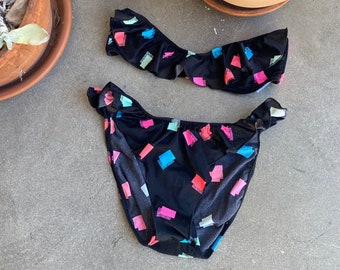 May include: Black bikini with a colorful geometric print. The top is strapless and has a ruffled edge. The bottom is a high-cut style with a ruffled edge.