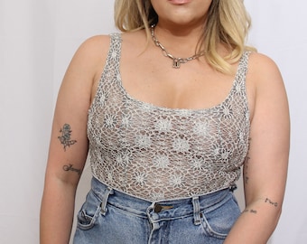 May include: A silver crochet top with a scoop neck and thin straps. The top is sheer and has a floral pattern. The model is wearing blue jeans.