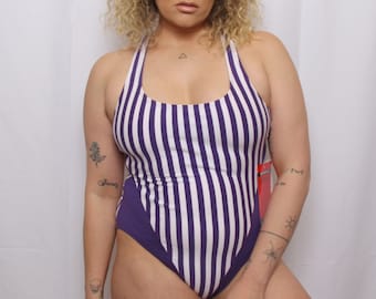 May include: A purple and white striped swimsuit with a halter neck and a high leg cut. The swimsuit has a solid purple panel on the bottom.