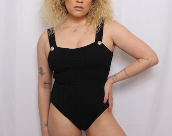 May include: A black one-piece swimsuit with silver buttons on the straps. The swimsuit has a square neckline and a fitted silhouette.