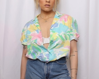May include: A woman wearing a short-sleeved, pastel-colored button-up shirt with a floral print. The shirt has a relaxed fit and is unbuttoned at the top. She is also wearing blue jeans.