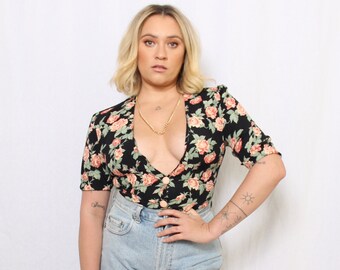 May include: A woman wearing a black floral print top with a plunging neckline and a gold chain necklace. She is also wearing light blue denim jeans.