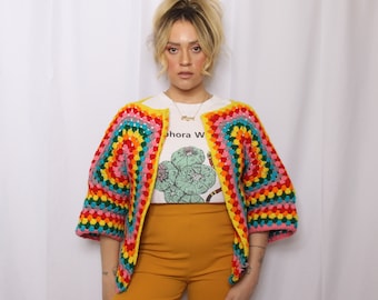 May include: A woman wearing a rainbow crochet cardigan with a white t-shirt that says "aphora W" and mustard yellow pants.