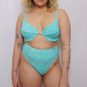 Puede incluir: Un bikini de color turquesa y blanco con estampado. La parte superior es de estilo halter con escote pronunciado y cierre de nudo en la espalda. La parte inferior es de estilo de cintura alta con un corte atrevido.