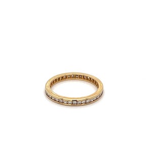 May include: A gold band ring with a continuous row of small, clear gemstones. The ring is set against a white background, highlighting its polished, reflective surface. The ring's design is simple and elegant.