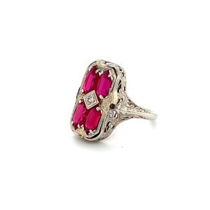 May include: An antique-style ring featuring a rectangular design with five vibrant red gemstones. A small, square diamond is set in the center. The ring has an ornate, filigree-style band and setting, with intricate detailing in the metalwork.