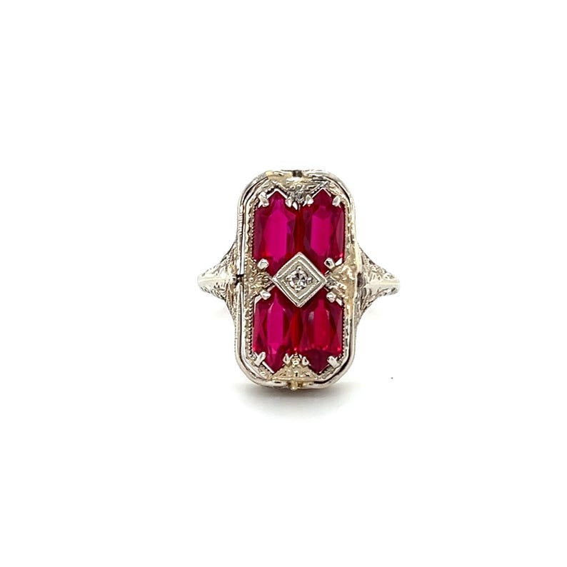 May include: A vintage-style ring with a rectangular face. The face features a central diamond surrounded by four rectangular, vibrant red gemstones. The ring has an ornate, filigree design on the band and setting, showcasing a classic, elegant aesthetic.