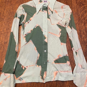 May include: A long-sleeved button-down shirt featuring a pointed collar and a geometric pattern in shades of sage green, pale mint, and soft peach. The fabric displays a dotted texture throughout, with a central button placket and cuffs.