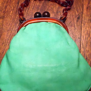 Peut inclure: Un sac à main vintage en cuir vert avec une bandoulière en plastique marron et deux accents en plastique marron sur le fermoir.