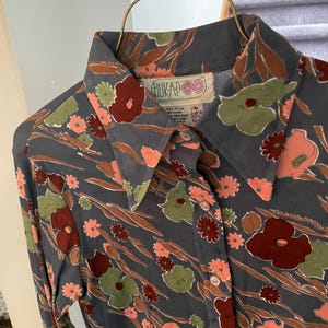 May include: A long-sleeve button-down shirt in a dark gray fabric featuring a floral pattern in shades of muted orange, deep red, and olive green. The shirt has a pointed collar and a label at the neckline that reads HUKAPOO with care instructions.