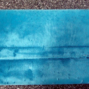 May include: Teal-colored clutch with a rectangular shape. The clutch features a fold-over flap and is accented with gold-toned metal details on the corners. The material appears to be a soft, velvety fabric.