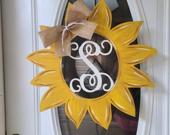 May include: A yellow sunflower door wreath with a burlap bow and a white monogram "S" in the center.