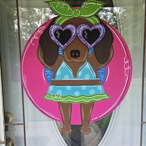 May include: A wooden door hanger featuring a cartoon dog wearing heart-shaped sunglasses, a blue polka-dot bikini, and a green bow. The dog is set against a pink circular background with white swirls.