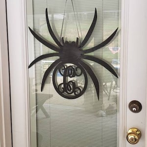 Puede incluir: Un colgador de puerta de araña de madera negra con un círculo monogramado en el centro. La araña está colgada de un gancho blanco en una puerta blanca.