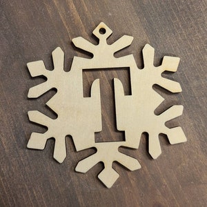May include: A wooden snowflake ornament with the letter "T" cut out in the center. The ornament is a light brown color and has a hole at the top for hanging.