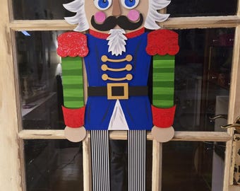 Puede incluir: Un colgador de puerta de madera de cascanueces con un esquema de color rojo, verde, azul y negro. El cascanueces tiene una barba blanca, un bigote negro y un ribete dorado en el sombrero.