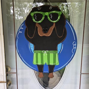 May include: A wooden door hanger featuring a dachshund wearing green sunglasses and swim trunks, floating in a blue inner tube. The dog is black with a brown snout and paws. The inner tube has a white outline.