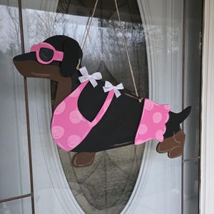 May include: A black dachshund wearing pink sunglasses and a pink polka dot swimsuit. The dachshund is hanging from a string and is a wooden cutout.