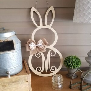 May include: A wooden bunny door hanger with a burlap bow and the letter "M" in a decorative font. The hanger is painted in a light wood color and is hanging on a wall.