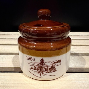 May include: A ceramic jar with a removable lid, featuring a two-tone brown and cream glaze. The lower cream section displays a brown printed illustration of a building and the year 1986. The upper section and lid are a glossy dark brown.