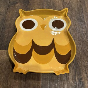 May include: A ceramic tray shaped like an owl, featuring a glossy yellow finish with large white and brown eyes, a small triangular beak, and dark brown scalloped wing details along the bottom edge, creating a distinct avian silhouette.