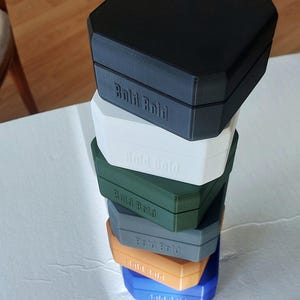 Può includere: Una pila di sei scatole esagonali in vari colori: nero, bianco, verde, grigio, arancione e blu. Ogni scatola ha il testo "Bald Bold" stampato. Le scatole sono impilate l'una sull'altra.