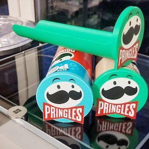 Könnte beinhalten: Ein grüner Pringles-Chip-Spender mit passendem Deckel und einer blauen Pringles-Dose. Der Spender hat das Pringles-Logo und ein Schnurrbart-Design. Die Dose ist rot, weiß und blau.