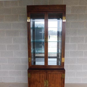 May include: A dark wood cabinet with glass doors and brass hardware. The cabinet has two shelves and a lower cabinet with two doors. The cabinet is in front of a gray brick wall.
