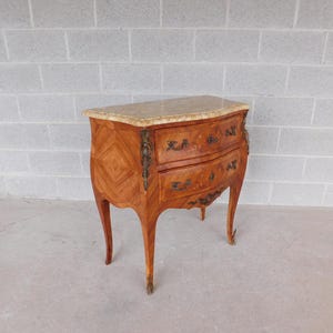 May include: A small, antique wooden chest of drawers with a marble top. The chest has two drawers with ornate brass hardware and curved legs. The wood has a warm, reddish-brown tone with a patterned design.