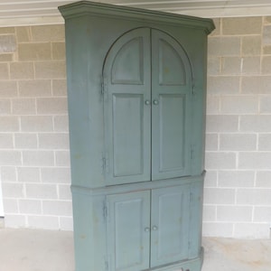 May include: A green corner cabinet with two sets of double doors. The top doors have arched tops and the bottom doors are rectangular. The cabinet has a simple design and is made of wood.