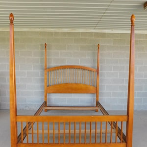May include: A wooden four-poster bed frame in a warm honey-toned finish. The bed features a headboard with vertical spindles and a footboard with a similar design. The tall posts are topped with decorative finials. The frame is made of wood.