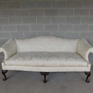 May include: Cream-colored sofa with a curved back and scrolled arms. The upholstery features a subtle floral pattern. The sofa has dark brown, carved wooden legs. The piece is set against a gray brick wall.
