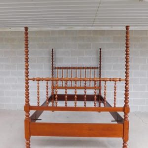 May include: A wooden four-poster bed frame in a warm, reddish-brown hue. The bed features turned posts and spindles, with a headboard and footboard. The frame is made of solid wood and has a classic, traditional design.
