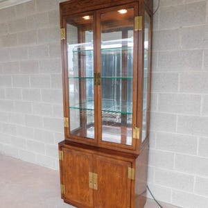 May include: A wooden display cabinet with glass doors and brass accents. The cabinet has two upper shelves and two lower doors. The cabinet is lit from within.