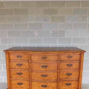 May include: A large, curved, wooden dresser with 12 drawers and ornate metal handles. The dresser is made of light wood with a warm, honey-colored finish.