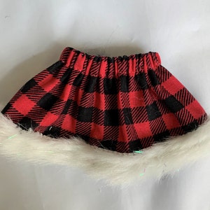 May include: A red and black plaid skirt with a white faux fur trim. The skirt has an elastic waistband. The fabric has a classic buffalo check pattern. This skirt is suitable for a doll or other small figure.