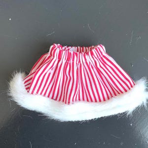 May include: A miniature skirt with red and white vertical stripes and a white faux fur trim along the bottom edge. The skirt is designed for a doll or small figure, and is likely part of a holiday or festive outfit.