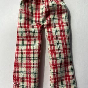 May include: Red, white, and green plaid pajama pants. The pants have an elastic waistband and a classic plaid pattern. The fabric appears to be a woven material. The pants are designed for a doll or small figure.