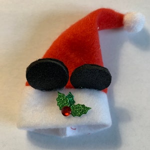 May include: A whimsical Santa hat ornament. The red felt hat has a white brim and a white pom-pom. Black, circular eyes and a green glitter holly with a red gem are attached to the white brim. A festive Christmas decoration.