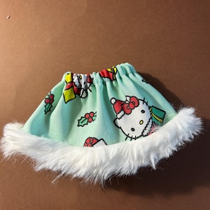 May include: A mint green skirt with a white faux fur trim. The skirt features a repeating pattern of Hello Kitty, holly, and wrapped gifts. The skirt is designed for a doll or small toy.