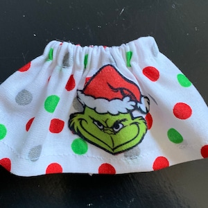 May include: A white fabric skirt with red, green, and gray polka dots. The skirt features an image of the Grinch wearing a Santa hat. The skirt is elasticized at the waist and is likely for a doll or small figure.