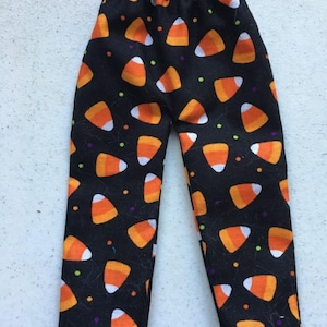 May include: A pair of black pants featuring an all-over print of candy corn pieces in orange, yellow, and white, accented with small scattered green and purple dots. The pants have an elasticized waistband and a straight-leg cut design.