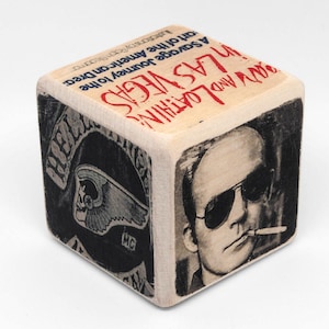 May include: A wooden cube featuring images and text from the novel "Fear and Loathing in Las Vegas." The cube displays a portrait of a man wearing sunglasses, the Hell's Angels logo, and the book's title in red letters. The cube is a literary-themed item.