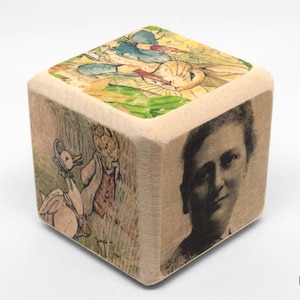 May include: A wooden cube with four different images on each side. One side features a black and white image of a woman, another side features a colorful illustration of a rabbit, a goose, and a flower, and another side features a colorful illustration of a goose and a flower.