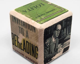 Puede incluir: Un cubo de madera con un diseño verde, amarillo y negro. El cubo tiene el texto "Invitation to a Book Reading" y "Lolita". El cubo también presenta una imagen en blanco y negro de un hombre con gafas.
