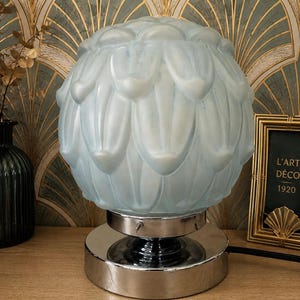 Peut inclure: Une lampe de table en verre bleu clair avec un abat-jour sphérique, ressemblant à une fleur, repose sur une base chromée. La lampe est de style Art Déco vintage. Une image encadrée avec le texte "L'ART DÉCO 1920" est visible.