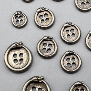 Puede incluir: Una colección de botones antiguos de color plateado. Cada botón presenta un diseño circular con cuatro agujeros y una serpiente que lo rodea. La cabeza de la serpiente está en la parte superior del botón.