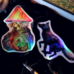 Puede incluir: Dos pegatinas holográficas con arte de fantasía. Una representa una figura con un sombrero de hongo y una cesta de plantas. La otra es una silueta de lobo llena de un diseño de galaxia. Las pegatinas están sobre una superficie negra.